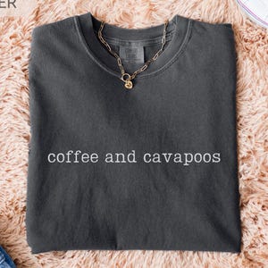 May include: A dark gray t-shirt with the words "coffee and cavapoos" in white. The shirt is folded and lies on a fluffy, peach-colored rug. A gold necklace and white sneakers are also visible.