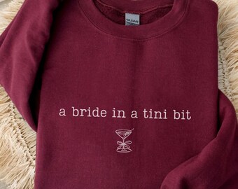 May include: Maroon sweatshirt with the text "a bride in a tini bit" and a cocktail graphic. The sweatshirt is displayed on a woven, circular mat. The word "MAROON" is printed at the bottom of the image.