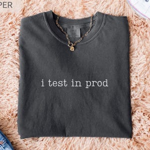 May include: A dark gray t-shirt with the text "i test in prod" in white. A gold chain necklace with a pendant rests on the shirt. The shirt is folded on a peach-colored, fluffy surface, with blue jeans and white sneakers visible.