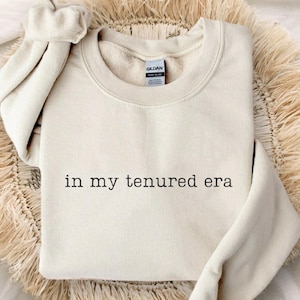 May include: A sand-colored sweatshirt with the phrase "in my tenured era" printed in black. The sweatshirt is displayed on a woven, circular placemat, with the sleeves rolled up. The word "SAND" is printed in a small, rounded rectangle.
