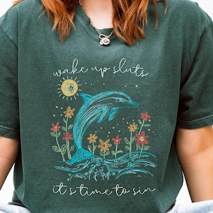 May include: A dark teal t-shirt with a graphic of a dolphin leaping out of the water, surrounded by flowers and the text "wake up sluts, it's time to sin". The shirt is labeled "Blue Spruce".