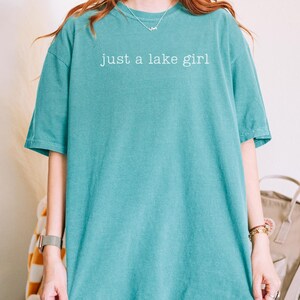 May include: Seafoam green t-shirt with the text "just a lake girl" in white. The shirt is a solid color and has short sleeves. The model is wearing an XL size.