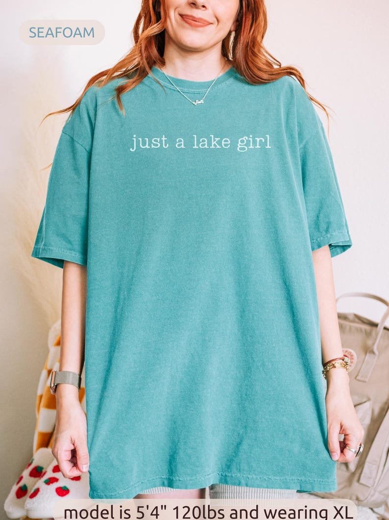 May include: Seafoam green t-shirt with the text "just a lake girl" in white. The shirt is a solid color and has short sleeves. The model is wearing an XL size.