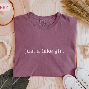 May include: Berry-colored t-shirt with the text "just a lake girl" in white. The shirt is surrounded by a white headband, a beige hat, a scrunchie, and white sneakers.