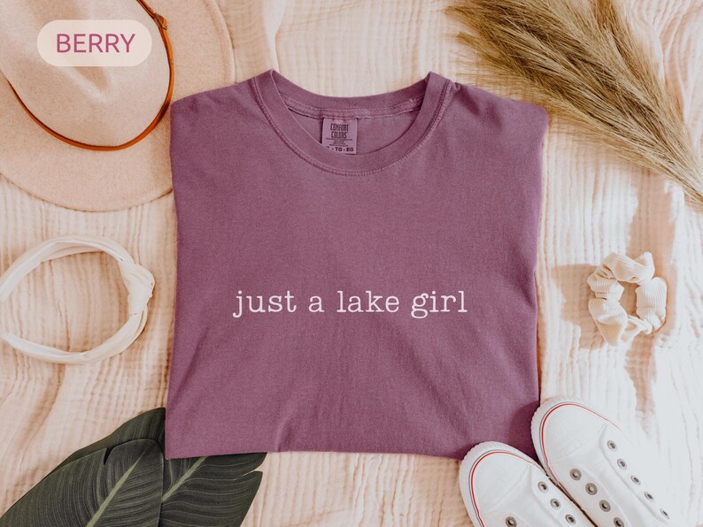 May include: Berry-colored t-shirt with the text "just a lake girl" in white. The shirt is surrounded by a white headband, a beige hat, a scrunchie, and white sneakers.