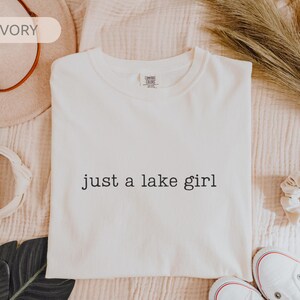 May include: Ivory-colored t-shirt with the text "just a lake girl" in black. The shirt is displayed on a textured, light-colored surface, with a hat, scrunchie, and shoes in the background.