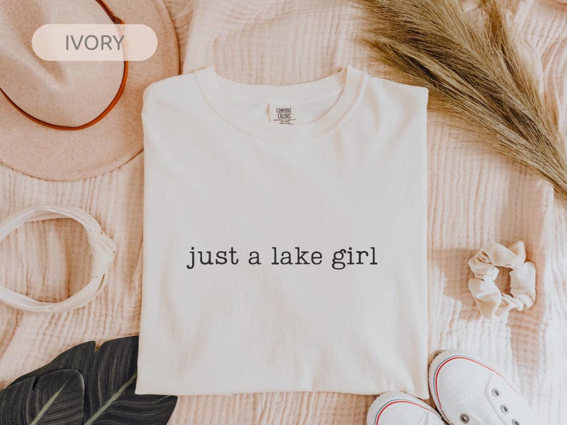 May include: Ivory-colored t-shirt with the text "just a lake girl" in black. The shirt is displayed on a textured, light-colored surface, with a hat, scrunchie, and shoes in the background.