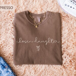 Puede incluir: Camiseta color espresso con las palabras "chosen daughter" en escritura cursiva y un peque&ntilde;o gr&aacute;fico de lazo. Tambi&eacute;n se ven un collar de cadena dorada y zapatillas blancas. La camiseta se muestra sobre una superficie esponjosa de color melocot&oacute;n.