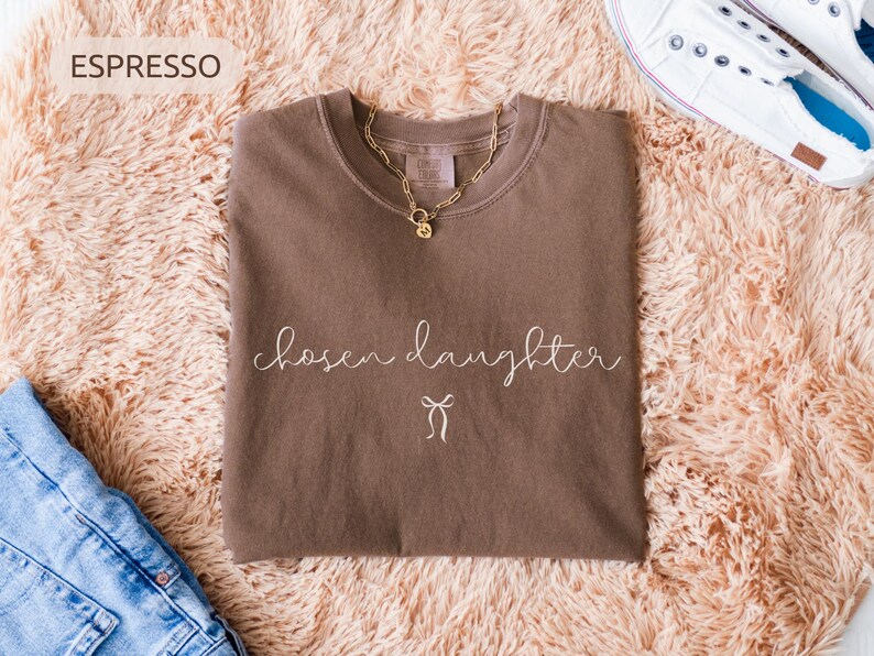 Puede incluir: Camiseta color espresso con las palabras "chosen daughter" en escritura cursiva y un peque&ntilde;o gr&aacute;fico de lazo. Tambi&eacute;n se ven un collar de cadena dorada y zapatillas blancas. La camiseta se muestra sobre una superficie esponjosa de color melocot&oacute;n.