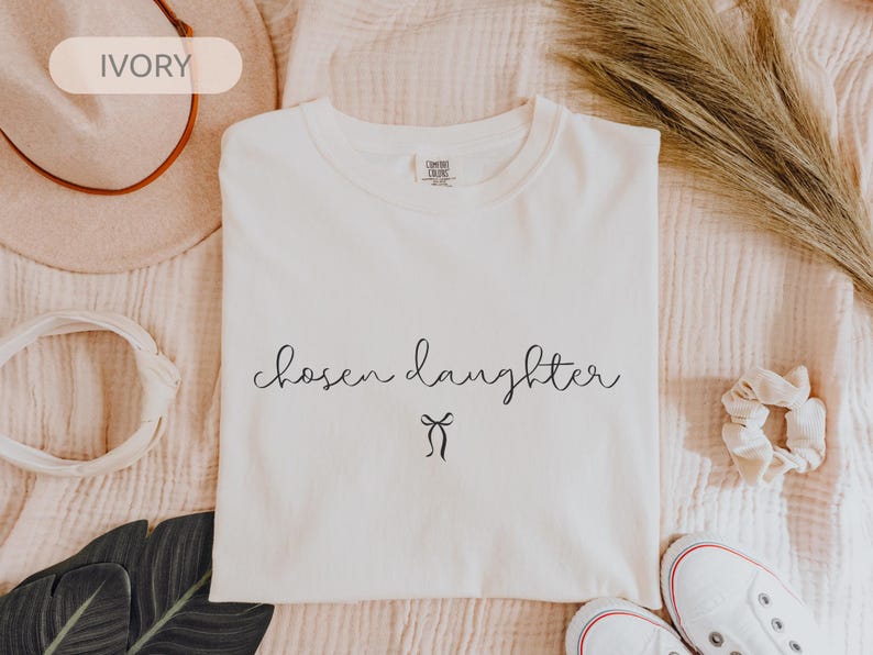 Puede incluir: Camiseta de color marfil con las palabras "chosen daughter" en fuente cursiva y un peque&ntilde;o lazo. La camiseta est&aacute; colocada plana con un sombrero, una goma para el pelo y zapatos en el fondo.