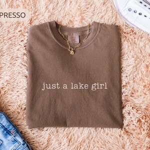 May include: Espresso-colored t-shirt with the text "just a lake girl" in white. A gold chain necklace with a pendant rests on the shirt. The shirt is displayed on a fluffy, peach-colored rug, with blue jeans and white sneakers.