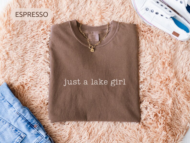 May include: Espresso-colored t-shirt with the text "just a lake girl" in white. A gold chain necklace with a pendant rests on the shirt. The shirt is displayed on a fluffy, peach-colored rug, with blue jeans and white sneakers.