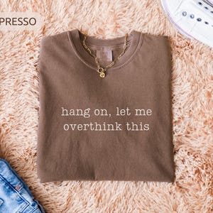 May include: A brown t-shirt with the text "hang on, let me overthink this" in white. A gold chain necklace is on the shirt. The shirt is displayed with blue jeans and white trainers.