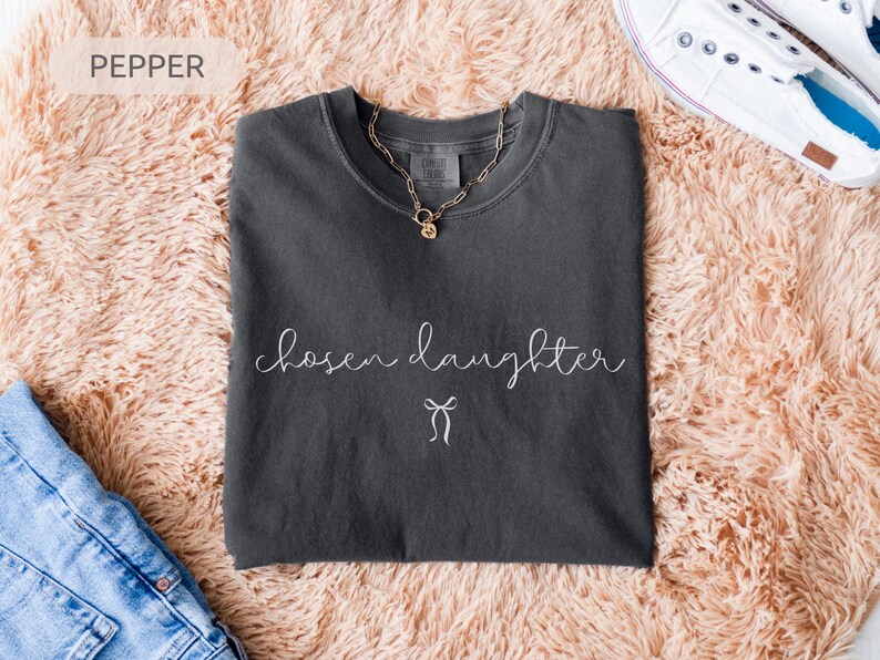 Puede incluir: Una camiseta gris oscuro con las palabras "chosen daughter" en escritura cursiva y un gr&aacute;fico de lazo. Un collar dorado descansa sobre la camiseta. La camiseta se muestra sobre una superficie melocot&oacute;n y esponjosa, con vaqueros azules y zapatillas blancas.