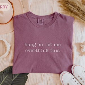 May include: Berry-coloured t-shirt with the phrase "hang on, let me overthink this" in white lettering. The shirt is surrounded by a white hat, scrunchie, and white trainers. A sprig of dried grass and a green leaf are also visible.
