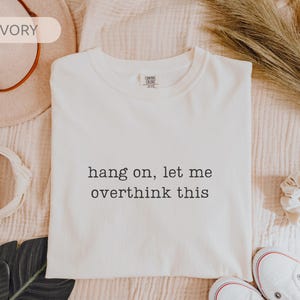 May include: Cream-coloured t-shirt with the text "hang on, let me overthink this" in black. The shirt is displayed with a hat, scrunchie, and shoes. A relaxed, everyday style.