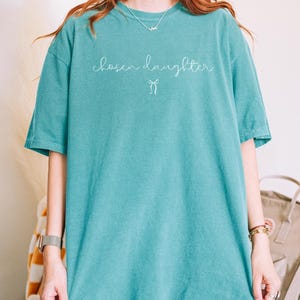 Puede incluir: Camiseta verde mar con la frase "chosen daughter" en cursiva y un peque&ntilde;o lazo. La camiseta es de corte holgado y tiene mangas cortas. La modelo lleva una talla XL.