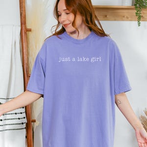 May include: A violet-colored t-shirt with the text "just a lake girl" in white. The shirt is a crew neck and has short sleeves. The word "VIOLET" is in a light purple box in the upper left corner.