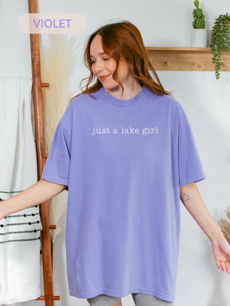 May include: A violet-colored t-shirt with the text "just a lake girl" in white. The shirt is a crew neck and has short sleeves. The word "VIOLET" is in a light purple box in the upper left corner.