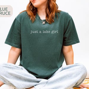 May include: A dark teal t-shirt with the text "just a lake girl" in white. The shirt is a relaxed fit and is paired with gray sweatpants. The words "Blue Spruce" are in a light beige box in the upper left corner.