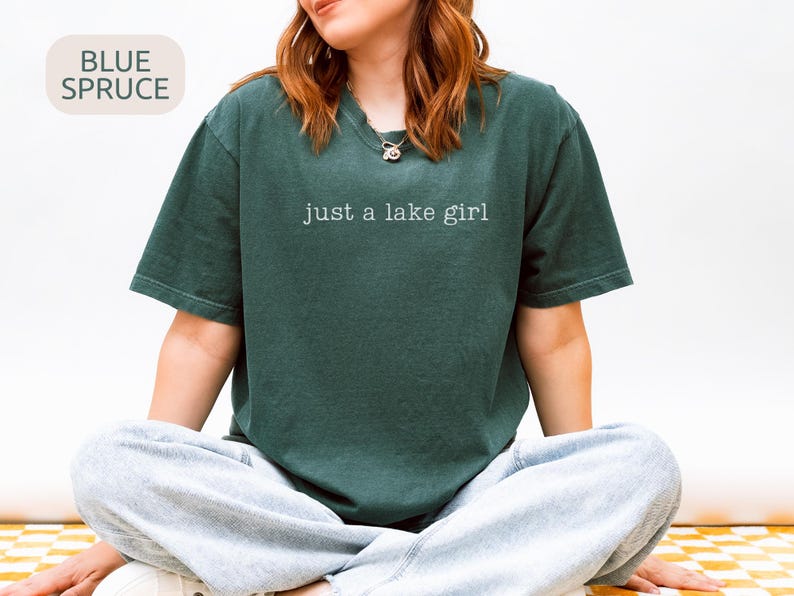 May include: A dark teal t-shirt with the text "just a lake girl" in white. The shirt is a relaxed fit and is paired with gray sweatpants. The words "Blue Spruce" are in a light beige box in the upper left corner.