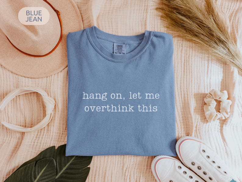 May include: A blue jean coloured t-shirt with the text "hang on, let me overthink this" in white. The shirt is folded and surrounded by a hat, scrunchie, shoes, and a leaf.
