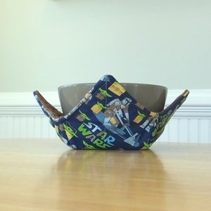 May include: A blue and green fabric bowl cozy with a Star Wars theme. The cozy is holding a brown ceramic bowl.