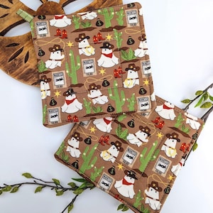 May include: Two brown square pot holders with a cowboy-themed pattern. The pattern includes cartoon ghosts wearing cowboy hats, cacti, wanted posters, and money bags. Each pot holder has a loop for hanging.