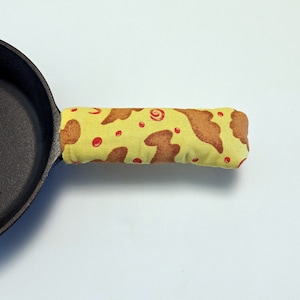 Puede incluir: Una sartén de hierro fundido negra con una cubierta de mango de tela amarilla y marrón. La cubierta del mango tiene puntos rojos y remolinos marrones.