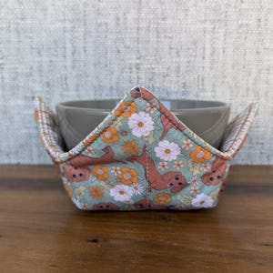May include: A green and brown fabric bowl cozy with a floral pattern and small animal illustrations. The cozy is designed to fit a standard-sized bowl.