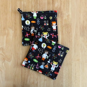 May include: Two black square pot holders with a science-themed pattern. The pattern includes cartoon scientists, atoms, beakers, and light bulbs in various colors. Each pot holder has a small loop for hanging.