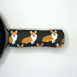 May include: A black cast iron skillet with a dark gray fabric handle cover. The handle cover has a repeating pattern of brown and white corgi dogs.
