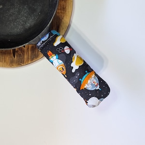 May include: A black fabric handle cover with a space-themed pattern featuring planets, rockets, and fried eggs. The cover is designed to fit over a pan handle, providing a heat-resistant grip. The pan is black and sits on a wooden surface.