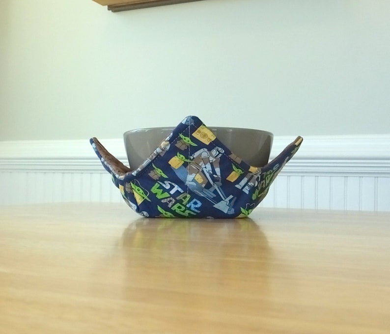 May include: A blue and green fabric bowl cozy with a Star Wars theme. The cozy is holding a brown ceramic bowl.