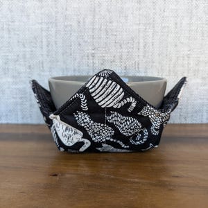 May include: A black and white patterned bowl cozy with a gray bowl inside. The cozy is made of fabric and has a white and black design of abstract shapes.