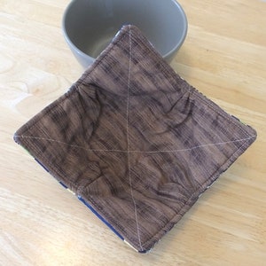 May include: A brown quilted bowl cozy with a woodgrain pattern. The cozy is made of fabric and has a square shape.