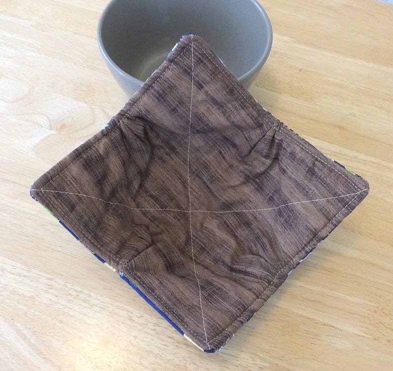 May include: A brown quilted bowl cozy with a woodgrain pattern. The cozy is made of fabric and has a square shape.