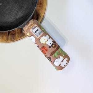 May include: A brown pot handle cover with a repeating pattern of chef characters, cowboy hats, and the word "WANTED". The cover is placed on a black cast iron skillet and a wooden trivet.