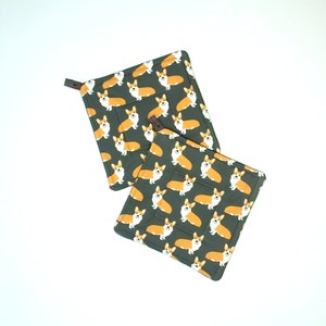 May include: Two square pot holders with a dark green background and a repeating pattern of orange and white corgi dogs. Each pot holder has a small brown loop for hanging.