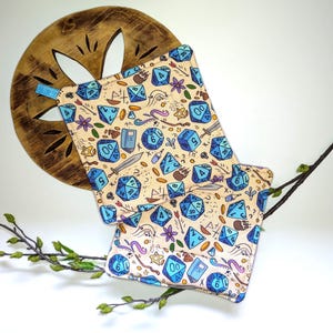 May include: Two beige pot holders with a blue dice pattern, a blue loop, and a wooden decorative piece. The pot holders feature blue dice, swords, books, and other fantasy-themed illustrations. The background is white.