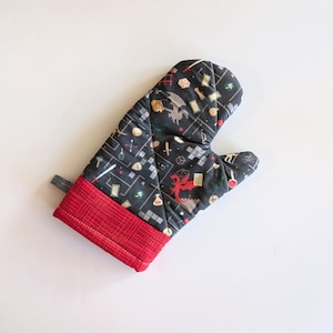 以下が含まれることがあります： 赤い袖口が付いた黒いオーブンミトン。ミトンは、ドラゴン、サイコロ、ポーション、その他のファンタジーをテーマにしたアイコンの繰り返しパターンを特徴としています。デザインはグリッドパターンに対して設定されています。