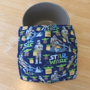 May include: A blue and green quilted bowl cozy with a Star Wars theme. The cozy features a repeating pattern of Mandalorian and Baby Yoda characters. The text "STAR WARS" is printed in white on the cozy.