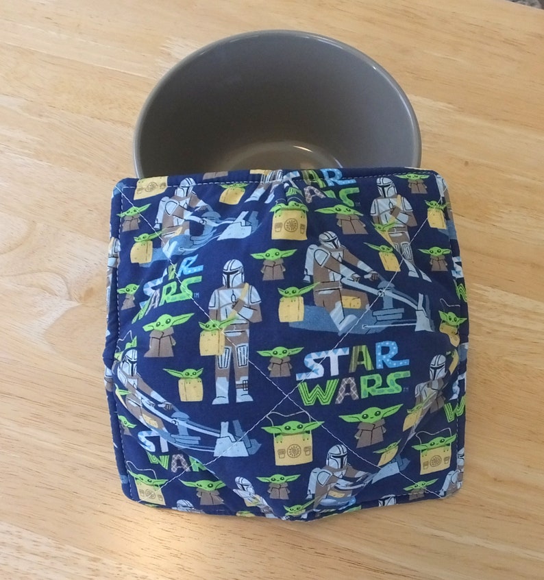 May include: A blue and green quilted bowl cozy with a Star Wars theme. The cozy features a repeating pattern of Mandalorian and Baby Yoda characters. The text "STAR WARS" is printed in white on the cozy.
