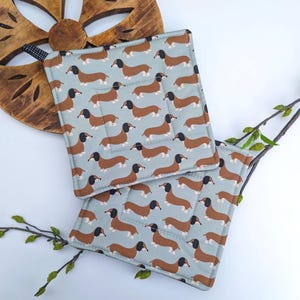 May include: Two square pot holders with a light blue background and a repeating pattern of brown dachshund dogs. The pot holders are quilted and have a brown border. A wooden trivet and greenery are in the background.