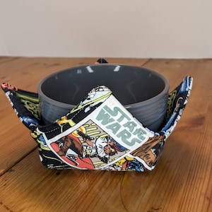 Może przedstawiać: Szara ceramiczna miska znajduje się w czarno-białym, materiałowym ocieplaczu do misek. Ocieplacz ma nadruk komiksu Star Wars z napisem "Star Wars".