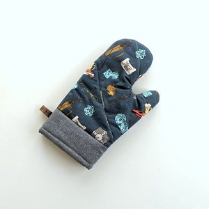 May include: A dark blue oven mitt with a gray cuff and a brown loop. The mitt is patterned with fantasy-themed illustrations, including dice, scrolls, and weapons. The mitt is designed for kitchen use.