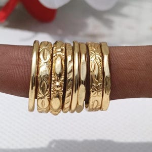 May include: A set of seven gold-toned rings with varying textures and patterns, including smooth bands, embossed oval shapes, and floral engravings, arranged side-by-side to showcase the distinct metallic surface details of each individual ring.