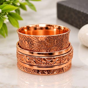 May include: A stack of three copper-colored metal rings featuring intricate embossed patterns. The top ring has a wide band with a floral-like texture, while the two lower rings display detailed geometric and repeating motifs in a metallic finish.