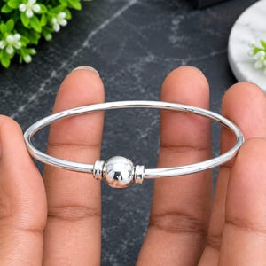 May include: A polished silver-tone bangle bracelet featuring a single, smooth, spherical bead at the center. The bracelet has a thin, rounded band design with a reflective metallic finish, showcasing a clean and simple aesthetic for wrist wear.