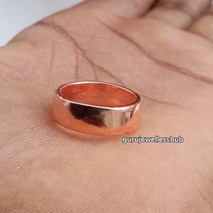 Könnte beinhalten: Ein massiver Kupferring mit glatter, polierter Oberfläche und flachem Bandprofil. Das Metall hat einen warmen, rötlich-orangen metallischen Glanz. Der Text gurujewellershub ist in der unteren rechten Ecke des Bildes sichtbar.
